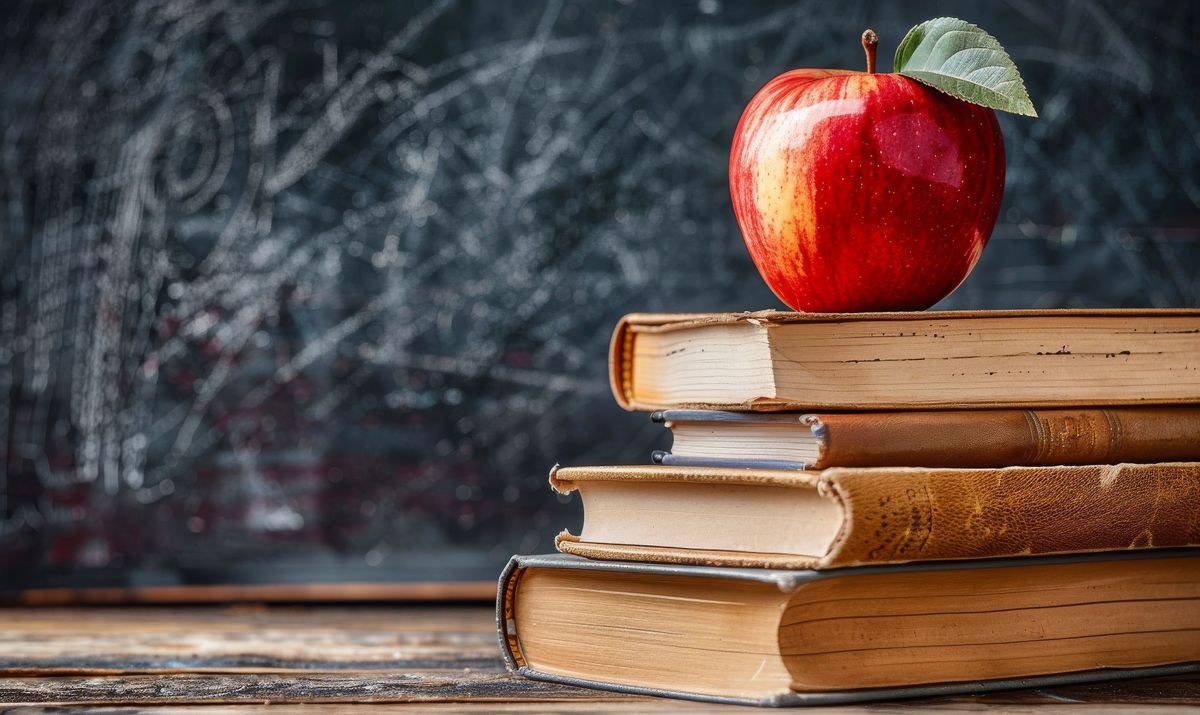 Stack of well-worn books with a red apple in front of a chalkboard